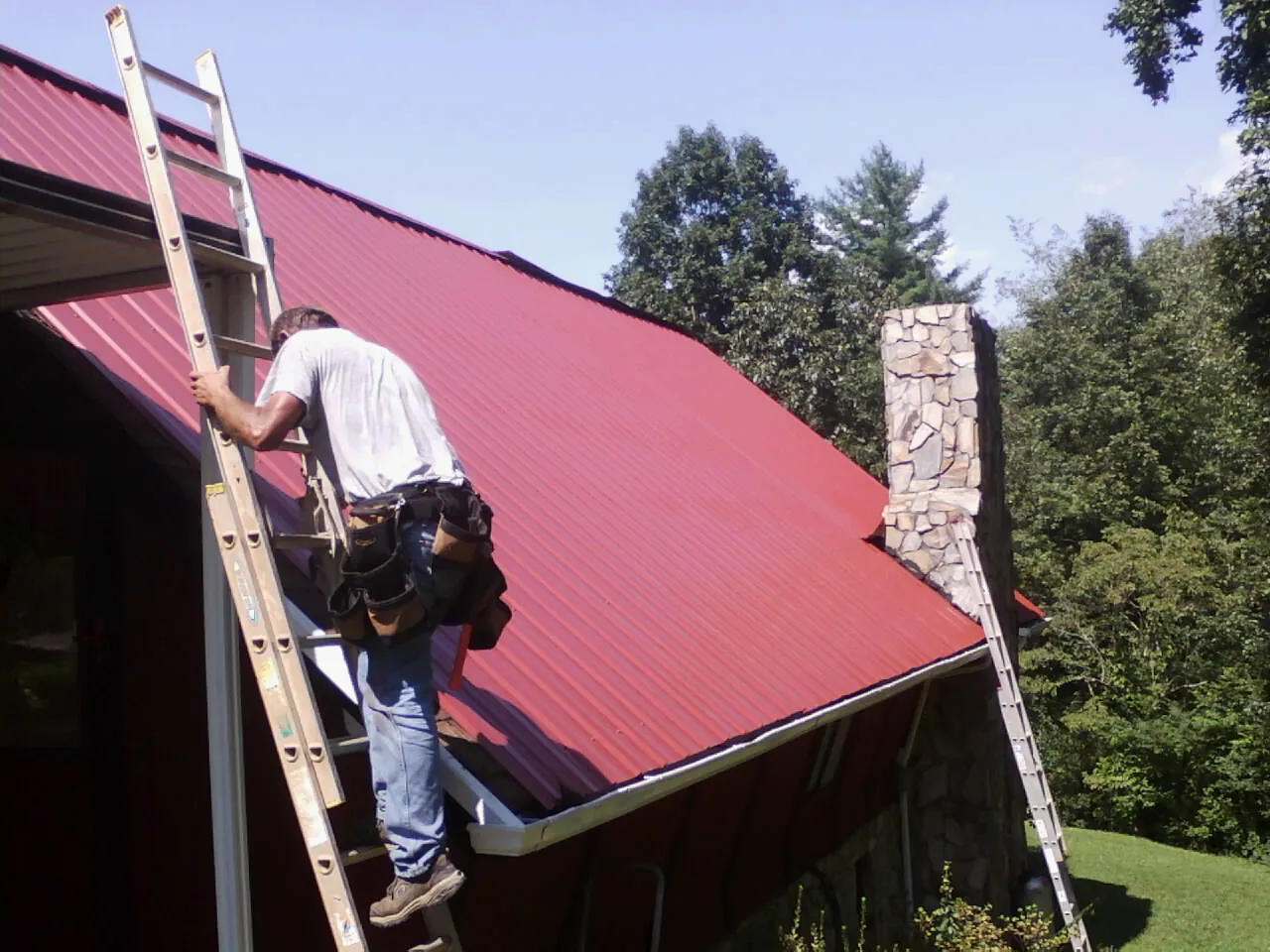 Roofer on a ladder beside a red metal roof and stone chimney — Soffit & Fascia Repair by Roofing Brothers in Lake Hamilton